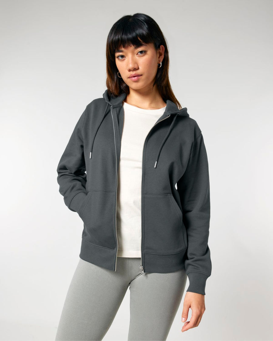 A woman stands facing the camera, wearing a dark grey Stanley/Stella STSU179 Stella/Stella Cultivator 2.0 The Iconic Unisex Zip-thru Hoodie Sweatshirt over a light beige shirt, with light grey pants, against a plain background.