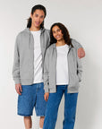Two individuals stand together, both wearing grey Stanley/Stella STSU179 Stella/Stella Cultivator 2.0 The Iconic Unisex Zip-thru Hoodie Sweatshirts made from brushed fleece and organic cotton, white t-shirts, and blue jeans. One has an arm around the other's shoulder. They are smiling in front of a plain background.