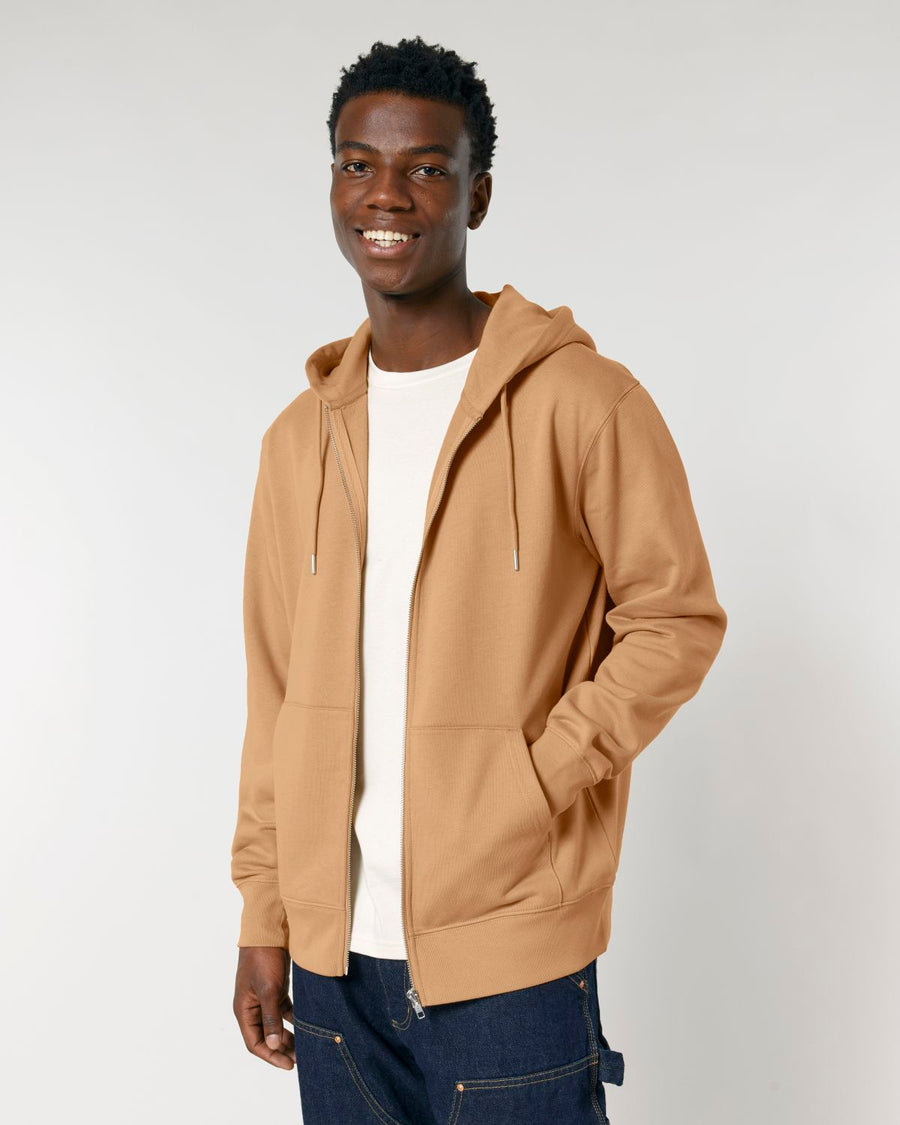 A person in a tan Stanley/Stella STSU179 Stella/Stella Cultivator 2.0 The Iconic Unisex Zip-thru Hoodie Sweatshirt made from organic cotton and a white t-shirt stands smiling against a plain background.