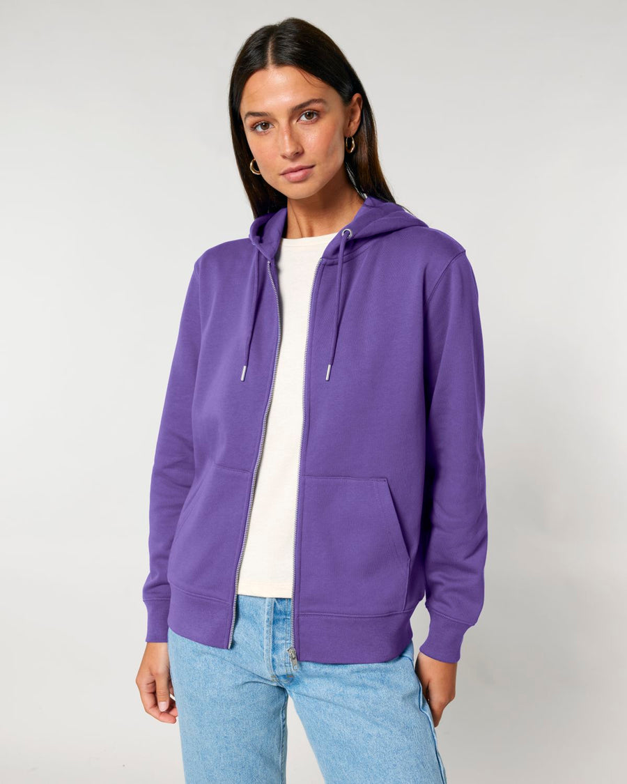 A person with long dark hair is wearing a purple STSU179 Stella/Stella Cultivator 2.0 The Iconic Unisex Zip-thru Hoodie Sweatshirt by Stanley/Stella over a white shirt and light blue jeans, standing against a plain background.