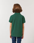 A child with medium-length brown hair is seen from the back, wearing the Stanley/Stella STPK908 Mini Sprinter The Iconic Kids Polo in soft organic cotton and blue jeans against a plain backdrop.