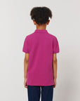 A child with curly hair faces away, wearing the Stanley/Stella STPK908 Mini Sprinter The Iconic Kids Polo in magenta organic cotton piqué and dark pants, set against a plain light background.