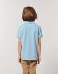 A child with medium-length brown hair, wearing the Stanley/Stella STPK908 Mini Sprinter The Iconic Kids Polo in light blue and beige pants, stands facing away from the camera against a plain background.