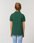 A child with braided hair, seen from behind, wears a Stanley/Stella STPK908 Mini Sprinter The Iconic Kids Polo in green and light blue jeans against a plain light background.