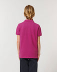 A person with light brown hair in a low braid is shown from behind, wearing the Stanley/Stella STPK908 Mini Sprinter The Iconic Kids Polo in magenta and dark pants against a plain background.