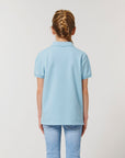 A girl with braided hair, facing away, wears the Stanley/Stella STPK908 Mini Sprinter The Iconic Kids Polo in light blue and matching jeans against a plain background.
