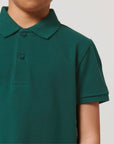 A child wears the STPK908 Stanley/Stella Mini Sprinter The Iconic Kids Polo in green, featuring short sleeves, a soft piqué fabric, a collar, and two buttons. The image displays the upper torso and part of the face.