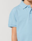 A child wears the STPK908 Stanley/Stella Mini Sprinter The Iconic Kids Polo in light blue, made from soft piqué organic cotton, shown from shoulders to upper arm against a plain background.