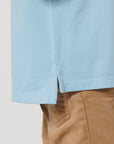 Close-up of a child wearing the Stanley/Stella STPK908 Mini Sprinter The Iconic Kids Polo in light blue, featuring piqué texture, a side slit at the hem, and tan pants with part of the pocket visible.