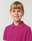 A young child with light brown hair and freckles smiles at the camera in a magenta Stanley/Stella STPK908 Mini Sprinter The Iconic Kids Polo, made from soft organic cotton piqué, set against a plain light background.