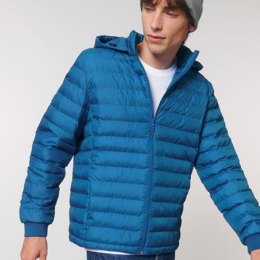 A person wearing a blue Stanley/Stella STJM837 Stanley Voyager Recycled Polyester Padded Jacket and a gray beanie looks down while slightly turning. The background is plain.