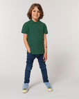 A child with medium-length brown hair wears the Stanley/Stella STPK908 Mini Sprinter The Iconic Kids Polo in green, blue jeans, and sneakers, standing against a plain light background with one hand in a pocket.