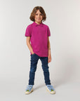 A child with long brown hair wears the Stanley/Stella STPK908 Mini Sprinter Kids Polo in magenta, paired with blue jeans and sneakers, standing against a plain white background.