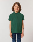 A child with shoulder-length brown hair wears a green Stanley/Stella STPK908 Mini Sprinter The Iconic Kids Polo in soft organic cotton piqué and blue jeans, standing facing forward against a plain light background.