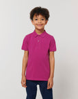 A child with curly hair wears a magenta STPK908 Stanley/Stella Mini Sprinter The Iconic Kids Polo and dark blue pants, standing against a plain light background.