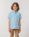 A young child with shoulder-length brown hair faces the camera, wearing a light blue Stanley/Stella STPK908 Mini Sprinter The Iconic Kids Polo and beige pants against a plain background.