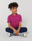 A young child with curly hair, dressed in a Stanley/Stella STPK908 Mini Sprinter The Iconic Kids Polo, blue jeans, and sneakers, sits cross-legged on the floor against a plain light background.