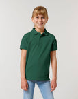 A young person smiles, wearing the Stanley/Stella STPK908 Mini Sprinter The Iconic Kids Polo in dark green organic cotton piqué and light blue jeans against a plain light background.
