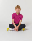 A young child with blonde hair sits cross-legged on the floor in a magenta Stanley/Stella STPK908 Mini Sprinter The Iconic Kids Polo, black pants, and yellow-soled white shoes, set against a plain white background.