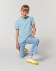 A young blonde girl kneels on one knee, wearing the Stanley/Stella STPK908 Mini Sprinter The Iconic Kids Polo in soft light blue organic cotton piqué, paired with light blue jeans and white sneakers with yellow soles against a plain white background.