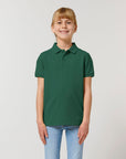 A young girl with blonde hair in a ponytail wears the Stanley/Stella STPK908 Mini Sprinter The Iconic Kids Polo in dark green, made from organic cotton piqué, paired with light blue jeans against a plain light background.