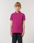 A young person with blond hair wears the Stanley/Stella Mini Sprinter The Iconic Kids Polo (STPK908) in magenta, crafted from soft organic cotton piqué, paired with black jeans and standing against a plain, light background.