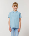 A young girl faces forward in the Stanley/Stella Mini Sprinter The Iconic Kids Polo (light blue, soft piqué organic cotton) with blue jeans, standing against a plain white background.