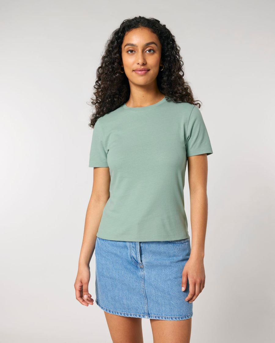 A woman with long curly hair wearing a STTW174 Stella Ella Womens Fitted Loose Knit T-Shirt by Stanley/Stella in green and a blue denim skirt stands against a plain background.