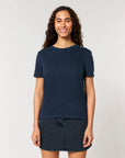 A woman with curly hair wearing a navy blue STTW174 Stella Ella Womens Fitted Loose Knit T-Shirt by Stanley/Stella and a denim skirt stands against a plain white background, smiling.