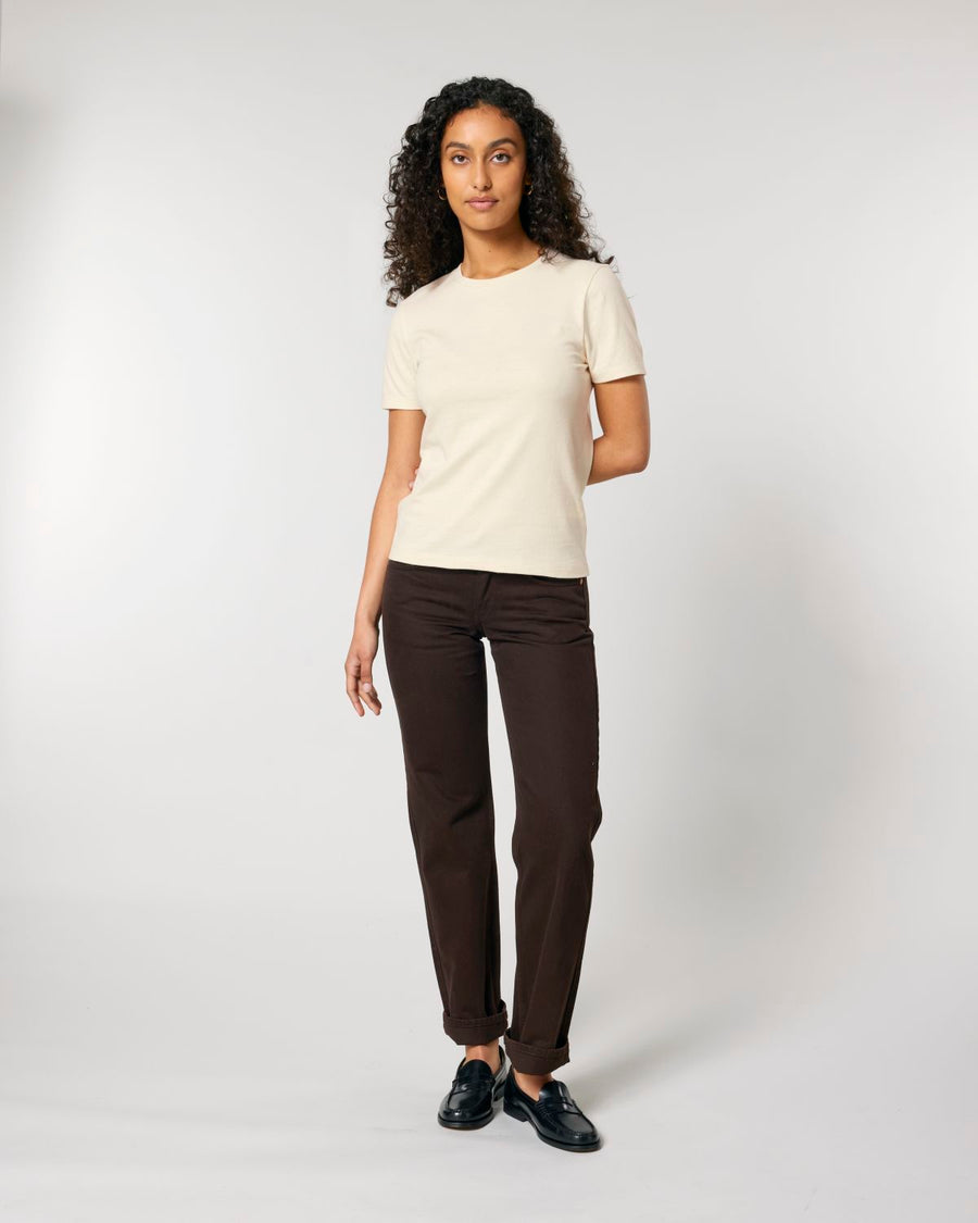 A person with curly hair wearing a white STTW174 Stella Ella Women's Fitted Loose Knit T-Shirt from Stanley/Stella, dark pants, and black shoes stands against a plain white background.