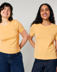 Two people are standing side by side, smiling, while wearing matching yellow STTW174 Stella Ella Womens Fitted Loose Knit T-Shirts by Stanley/Stella and blue jeans. The background is plain and light-colored.