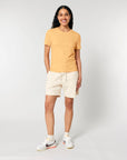 Person standing against a plain background, wearing a yellow STTW174 Stella Ella Womens Fitted Loose Knit T-Shirt by Stanley/Stella, light-colored shorts, white socks, and white sneakers with orange and black accents. Smiling and looking at the camera.