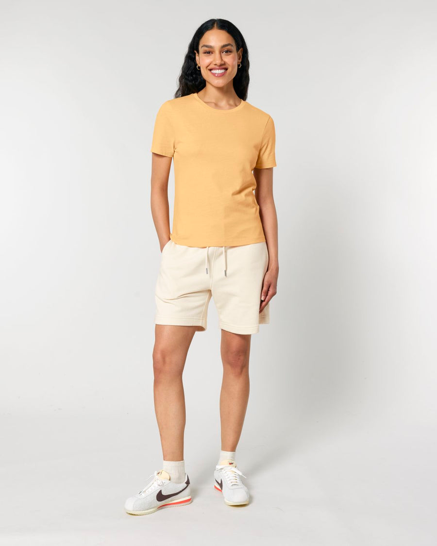 Person standing against a plain background, wearing a yellow STTW174 Stella Ella Womens Fitted Loose Knit T-Shirt by Stanley/Stella, light-colored shorts, white socks, and white sneakers with orange and black accents. Smiling and looking at the camera.