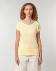 A woman with curly hair wears a light yellow STTW112 Stella Rounder Slub Rolled Sleeve T-Shirt by Stanley/Stella made from organic cotton and white pants, standing against a plain white background.
