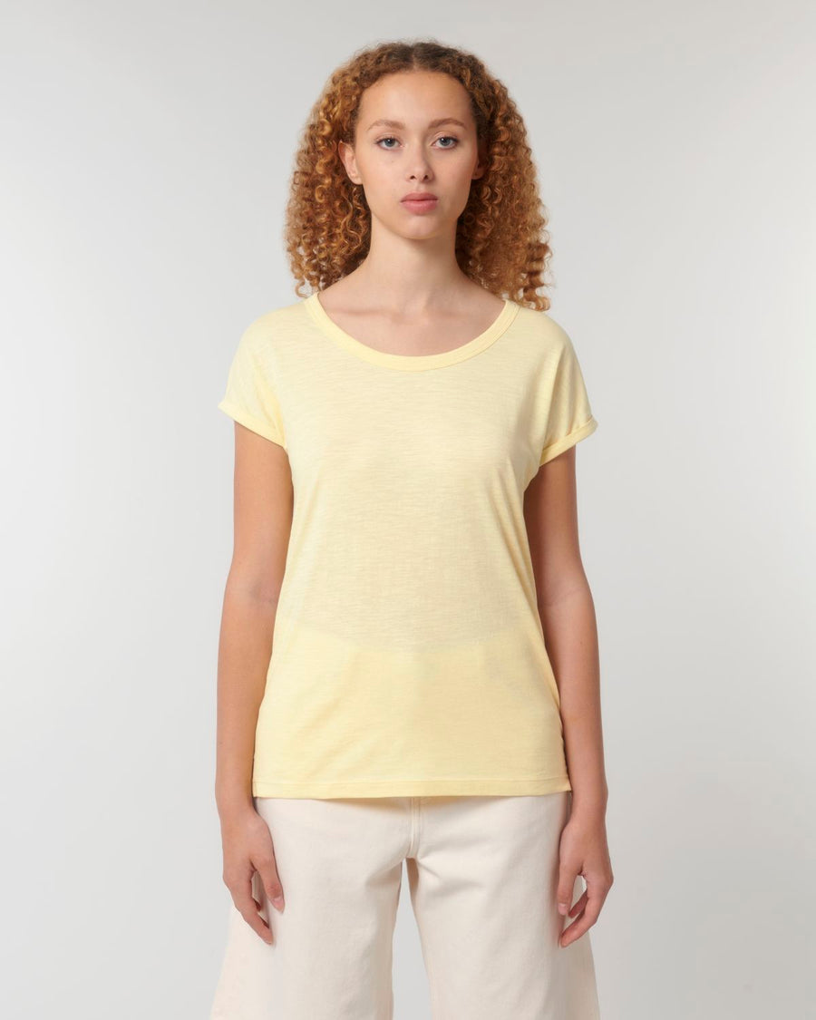 A woman with curly hair wears a light yellow STTW112 Stella Rounder Slub Rolled Sleeve T-Shirt by Stanley/Stella made from organic cotton and white pants, standing against a plain white background.