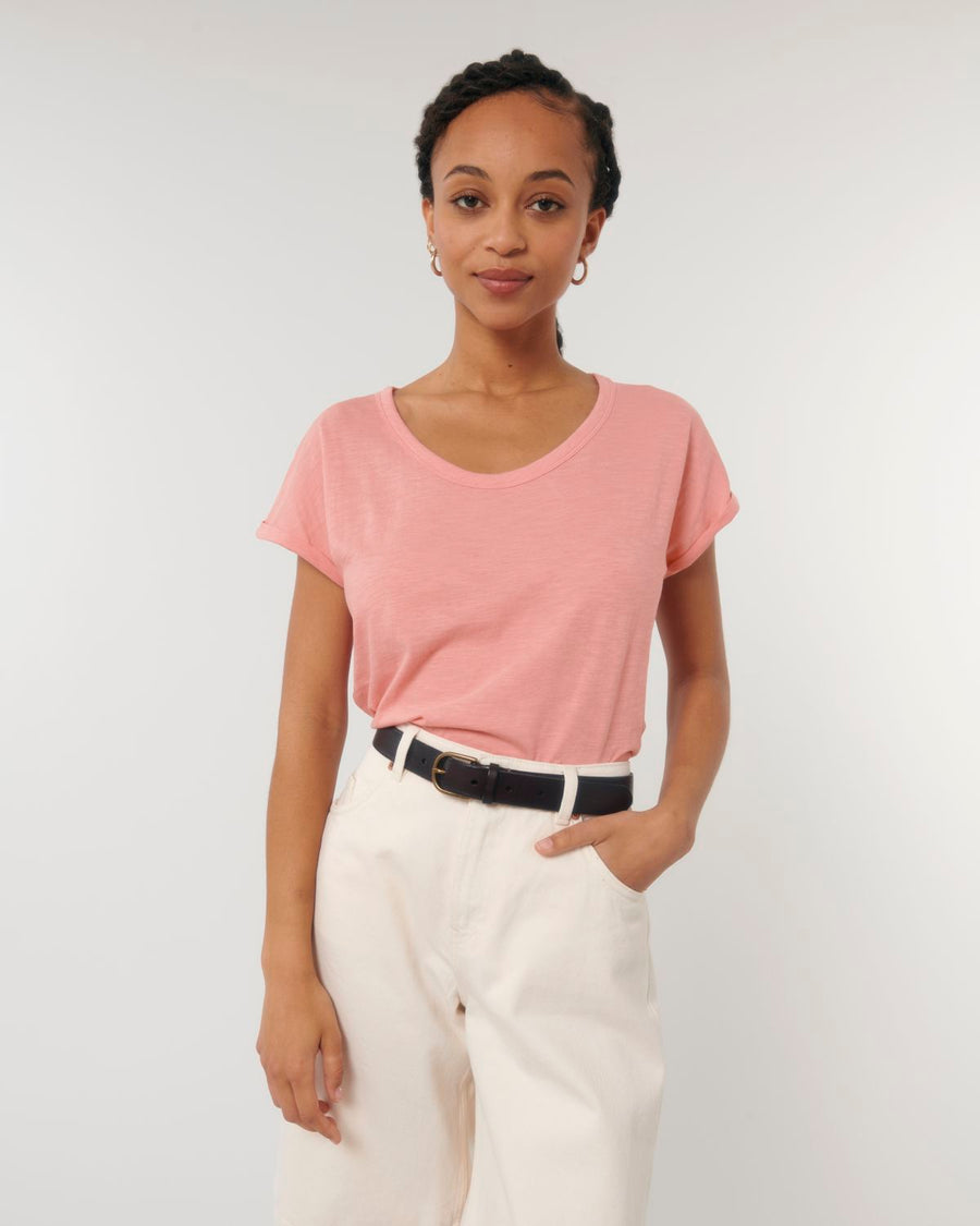 A person with braided hair stands against a plain background, wearing a pink Stanley/Stella STTW112 Stella Rounder Slub Rolled Sleeve T-Shirt tucked into high-waisted white pants, with one hand in their pocket.