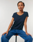 A person wearing a navy blue Stanley/Stella STTW112 Stella Rounder Slub Rolled Sleeve T-Shirt made of Organic Cotton and blue jeans sits on a stool against a plain white background.