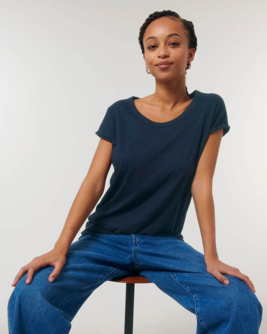 A person wearing a navy blue Stanley/Stella STTW112 Stella Rounder Slub Rolled Sleeve T-Shirt made of Organic Cotton and blue jeans sits on a stool against a plain white background.