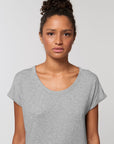 A woman with curly hair tied up, wearing a light gray STTW112 Stella Rounder Slub Rolled Sleeve T-Shirt by Stanley/Stella made from Organic Cotton, stands against a plain white background.