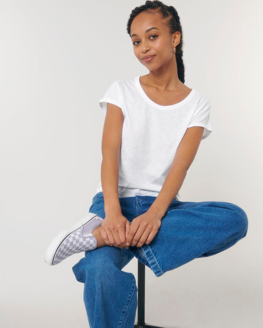 A person with braided hair sits on a stool, wearing a white Stanley/Stella STTW112 Stella Rounder Slub Rolled Sleeve T-Shirt made from organic cotton, blue jeans, and checkered slip-on sneakers. They have a slight smile and look towards the camera against a plain white background.