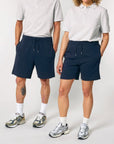 Two individuals standing side by side wearing light grey tops, STBU186 Stella/Stella Trainer 2.0 The Iconic Mid-light Unisex Jogger Shorts, white socks, and grey sneakers. The person on the left has a tattoo on their right leg.