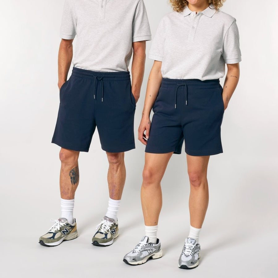 Two individuals standing side by side wearing light grey tops, STBU186 Stella/Stella Trainer 2.0 The Iconic Mid-light Unisex Jogger Shorts, white socks, and grey sneakers. The person on the left has a tattoo on their right leg.