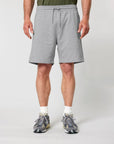 A person wearing a green shirt, white socks, and gray sneakers stands with their arms relaxed by their sides. They're dressed in gray Stanley/Stella STBU186 Stella/Stella Trainer 2.0 The Iconic Mid-light Unisex Jogger Shorts made from recycled polyester. The focus is on the lower half of their body.