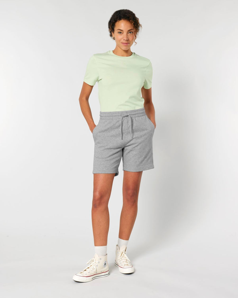 A woman stands against a plain background, wearing a light green T-shirt, STBU186 Stella/Stella Trainer 2.0 The Iconic Mid-light Unisex Jogger Shorts from Stanley/Stella, white socks, and white sneakers. She has her hands in her pockets and is smiling.