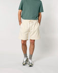 Person standing with hands in pockets, wearing a green T-shirt, beige organic cotton Stanley/Stella STBU186 Stella/Stella Trainer 2.0 The Iconic Mid-light Unisex Jogger Shorts, gray socks, and white sneakers against a plain background.