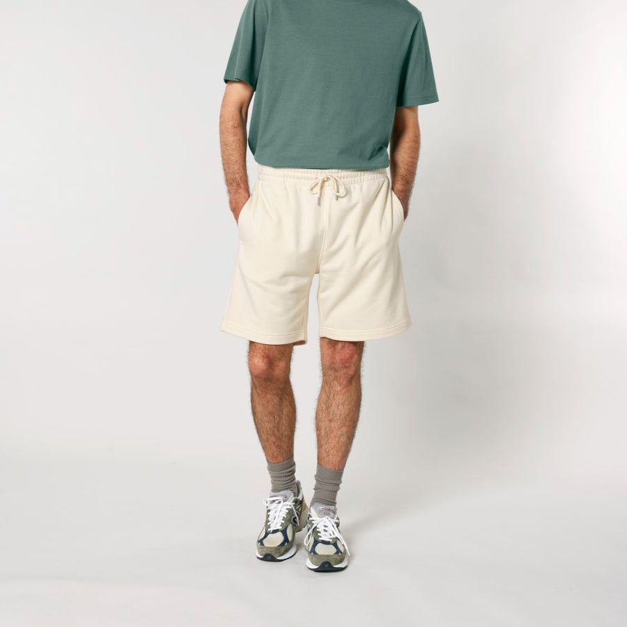 Person standing with hands in pockets, wearing a green T-shirt, beige organic cotton Stanley/Stella STBU186 Stella/Stella Trainer 2.0 The Iconic Mid-light Unisex Jogger Shorts, gray socks, and white sneakers against a plain background.