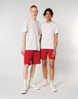 Two individuals stand against a plain background, both wearing light grey organic cotton t-shirts, Stanley/Stella STBU186 Stella/Stella Trainer 2.0 The Iconic Mid-light Unisex Jogger Shorts in red, and white high-top sneakers.