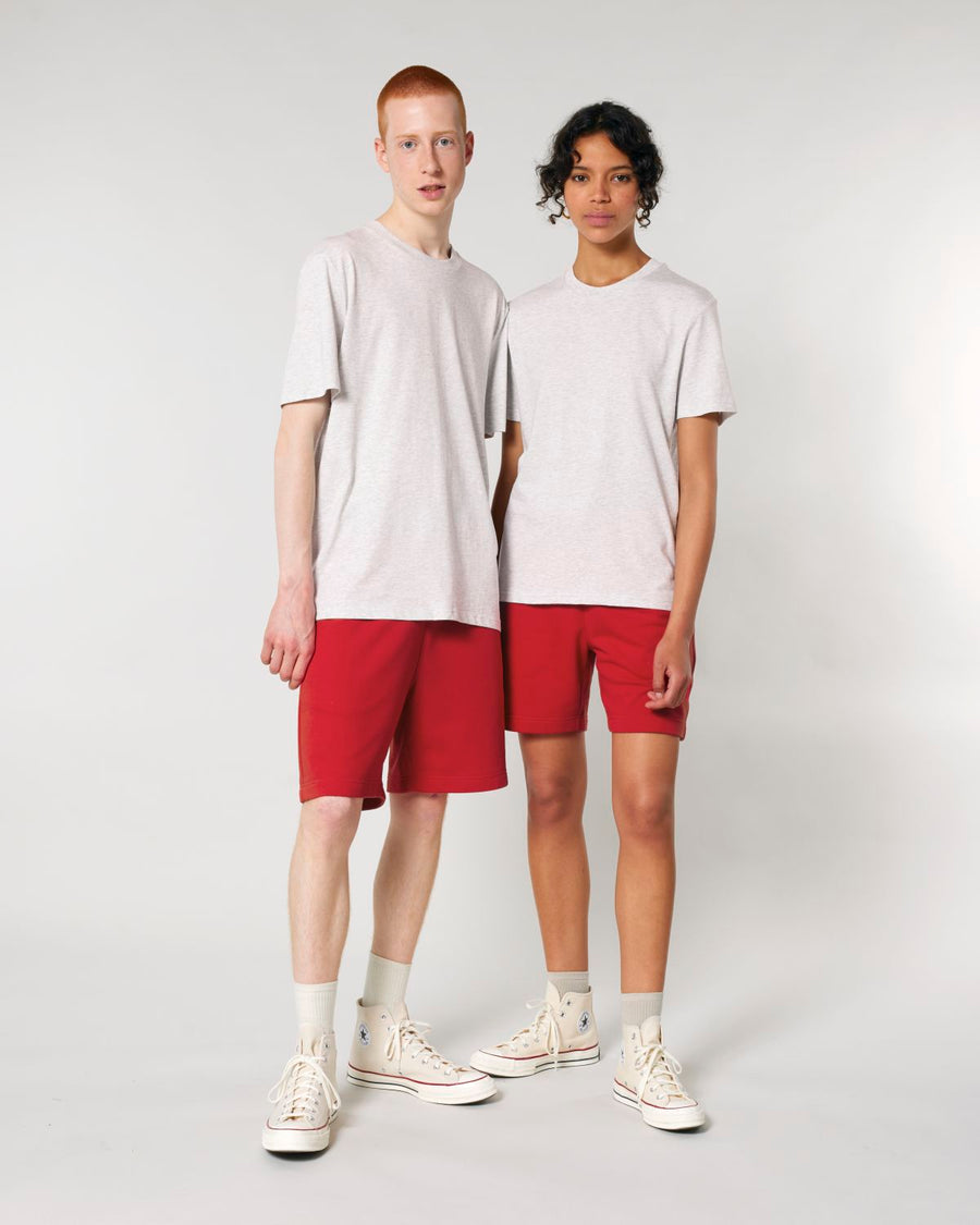 Two individuals stand against a plain background, both wearing light grey organic cotton t-shirts, Stanley/Stella STBU186 Stella/Stella Trainer 2.0 The Iconic Mid-light Unisex Jogger Shorts in red, and white high-top sneakers.