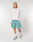 A person with long hair is standing against a plain background, wearing a light gray hoodie made from recycled polyester, Stanley/Stella STBU186 Stella/Stella Trainer 2.0 The Iconic Mid-light Unisex Jogger Shorts, white socks, and gray sneakers. Their hands are in the pocket of the hoodie.