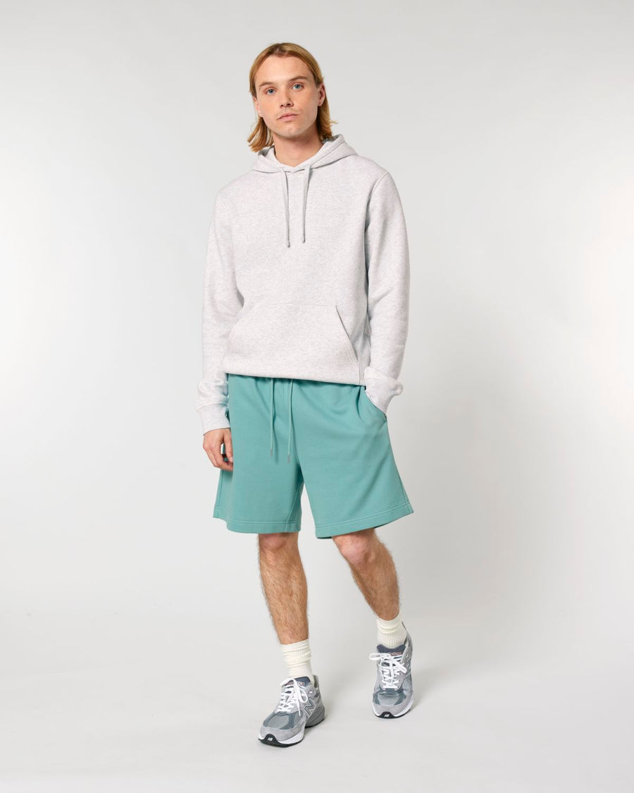 A person with long hair is standing against a plain background, wearing a light gray hoodie made from recycled polyester, Stanley/Stella STBU186 Stella/Stella Trainer 2.0 The Iconic Mid-light Unisex Jogger Shorts, white socks, and gray sneakers. Their hands are in the pocket of the hoodie.